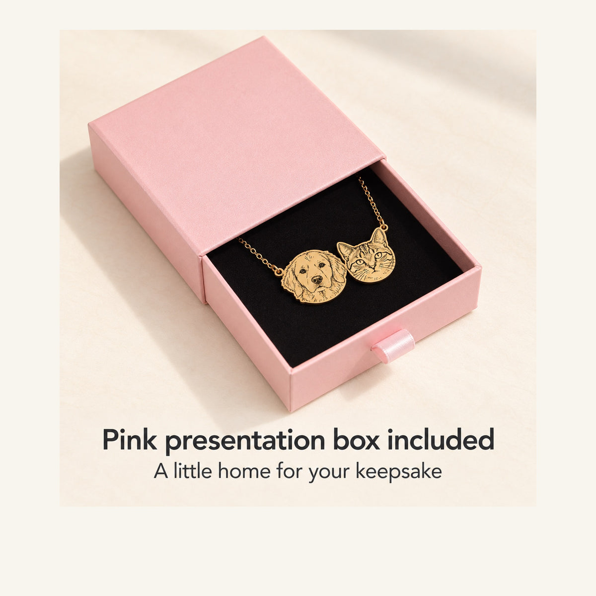 An open pink drawer box with a black insert holds a gold-colored necklace with connected retriever and tabby cat faces.